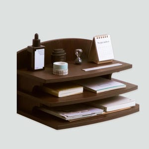 May include: A dark brown wooden desk organiser with three shelves. The top shelf holds a small calendar, an hourglass, and a bottle of serum. The lower shelves contain notebooks and papers. The organiser is designed for office or home use.