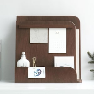 May include: A dark brown wooden desk organiser with a shelf, a small drawer, and a slot for holding papers. It features a white bottle, a gold paper clip, and several cards and papers. The organiser has a minimalist design.