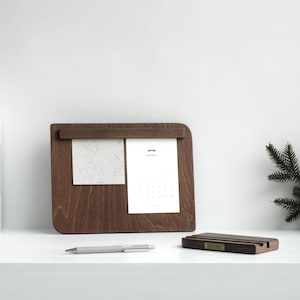 Puede incluir: Un organizador de escritorio de madera marrón oscuro con un calendario y una tarjeta de notas. El organizador tiene un soporte integrado para sostener un teléfono o una tableta. El organizador está sobre una superficie blanca.