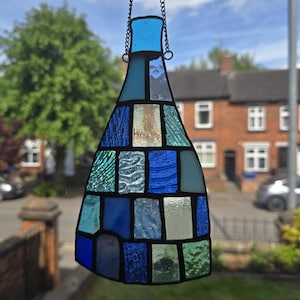 May include: A stained glass bottle-shaped suncatcher, suspended by a chain. It features a mosaic design of blue, teal, and clear glass pieces. The background shows a residential street with houses and trees, viewed through a window.