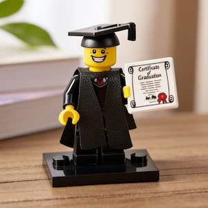 Könnte beinhalten: Eine Lego-Abschlussfigur mit schwarzer Kappe und Robe, die eine Urkunde mit der Aufschrift "Certificate of Graduation" hält. Die Figur hat einen gelben Kopf und steht auf einem schwarzen Sockel. Im Hintergrund sind Bücher und Grün zu sehen.