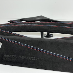 May include: Black interior trim pieces with red, white, and blue stitching. The trim has the text "M PERFORMANCE Limited edition" embossed on the surface. The pieces are curved and appear to be made of a suede-like material.