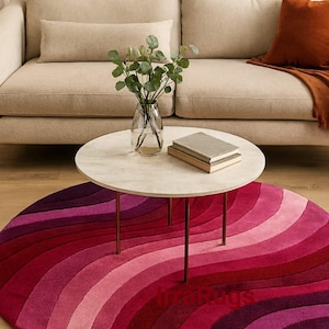 May include: A round rug with a gradient of red and pink hues, arranged in a wave pattern. The rug is placed beneath a marble-topped coffee table with brass legs, and a beige sofa. A vase of greenery and books are on the table.