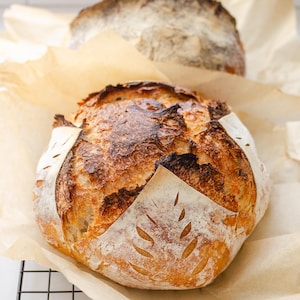 May include: Two round loaves of sourdough bread with a dark, golden-brown crust and a flour-dusted surface. The bread features a decorative wheat-stalk score pattern on the top and is resting on light brown parchment paper over a wire rack.