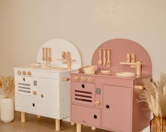 May include: Two wooden toy kitchens, one white and one pink, with wooden utensils and accessories. Each kitchen has a rounded top, a stove top, and a cabinet. The kitchens are on short, light-colored wooden legs.