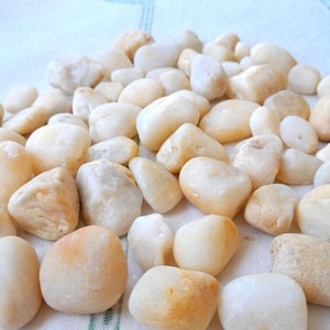 May include: A collection of smooth, rounded, cream-colored river rocks. The stones vary in size and shape, with some showing hints of tan and beige. They are arranged on a white surface, creating a natural, earthy aesthetic.