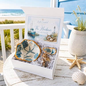 Op de afbeelding: Een witte geschenkdoos met The Harbour Collection van Irish Coastal Treasures, met een grote schelpschaal met blauwe zeester- en schelppatronen, een kleinere schelpschaal en een glazen flesje met schelpen, verpakt in witte papieren snippers.