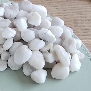 May include: A pile of smooth, white decorative stones. The stones vary in size and shape, with a rounded, polished appearance. They are arranged on a light green surface, suggesting a decorative or landscaping application.