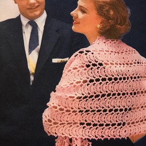 May include: A woman wearing a pink crocheted shawl with fringe. The shawl has a delicate pattern and is draped over her shoulders. She is standing next to a man wearing a dark suit and tie.