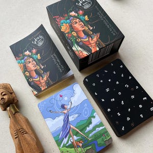 Könnte beinhalten: Ein Tarotkartenspiel mit einer schwarzen Schachtel und Kartenrücken mit weißen Himmelsymbolen. Die Schachtel und die Karten zeigen Illustrationen einer Frau mit floralem Kopfschmuck. Eine Karte zeigt eine Figur mit einem Hund, dem Text "O. The Fool".