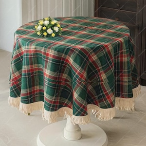 Puede incluir: Una mesa redonda cubierta con un mantel a cuadros verdes, rojos y crema. El mantel tiene un borde de flecos. Un pequeño ramo de flores blancas y amarillas está sobre la mesa. La mesa tiene una base de pedestal blanca.