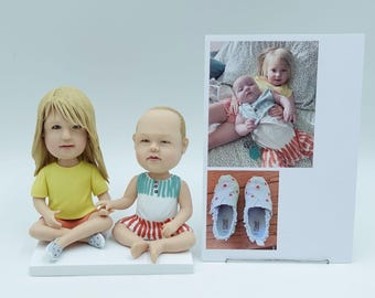 May include: Two bobblehead figurines of children, one with blonde hair in a yellow shirt and the other with a bald head in a white and blue shirt, sitting on a white base. A framed photo shows two children, and a pair of blue shoes with heart designs.