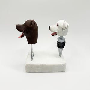 Puede incluir: Dos tapones de botella con temática de perros. Uno es una cabeza de perro marrón sobre un soporte de metal. El otro es una cabeza de perro blanca con manchas negras, unida a un tapón de botella negro y plateado. Ambos tienen lenguas rosas y se exhiben sobre una base blanca.