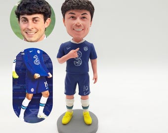 May include: A bobblehead figurine of a soccer player in a blue uniform with the number 3. The figurine has a smiling face and is standing on a gray base. The uniform includes blue shorts and yellow shoes.