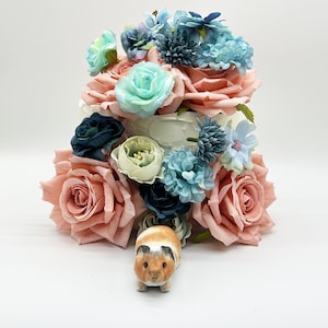 Peut inclure: Un g&acirc;teau d&eacute;coratif orn&eacute; de grandes roses couleur p&ecirc;che, de fleurs turquoise et bleues, et d'une petite figurine de cochon d'Inde &agrave; la base. Le g&acirc;teau a une base blanche et est d&eacute;cor&eacute; de divers arrangements floraux.