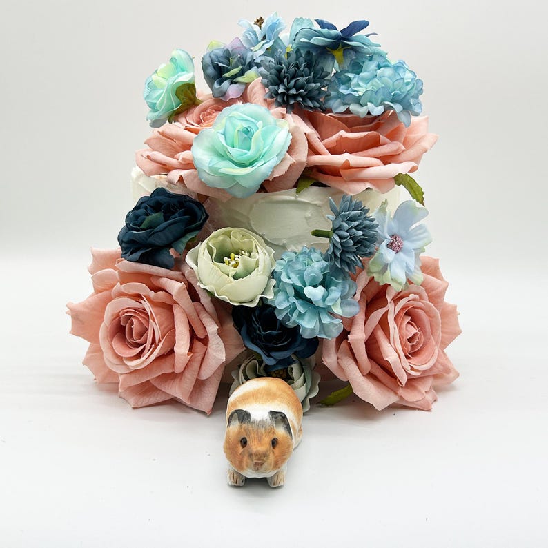 Peut inclure: Un g&acirc;teau d&eacute;coratif orn&eacute; de grandes roses couleur p&ecirc;che, de fleurs turquoise et bleues, et d'une petite figurine de cochon d'Inde &agrave; la base. Le g&acirc;teau a une base blanche et est d&eacute;cor&eacute; de divers arrangements floraux.