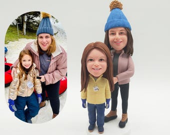 May include: A custom bobblehead figurine of a mother and daughter, both wearing blue knit hats. The mother is wearing a pink jacket and black pants, while the daughter is in a yellow jacket and blue pants. The image also includes a photo of the real people.