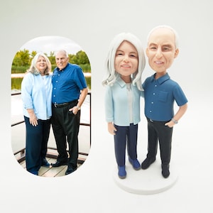 Könnte beinhalten: Eine personalisierte Wackelkopf-Figur eines Paares, mit einem Foto des Paares im Hintergrund. Die Figur zeigt einen Mann und eine Frau in Freizeitkleidung. Die Frau trägt ein hellblaues Hemd und der Mann trägt ein dunkelblaues Hemd.