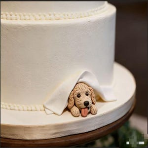 Könnte beinhalten: Eine mehrstöckige weiße Torte mit glattem Zuckerguss und einer Perlenbordüre. Ein kleiner, modellierter beigefarbener Welpe mit dunklen Augen und einer rosa Zunge schaut unter einer weißen Fondantdecke hervor, die am Tortenfuß drapiert ist.