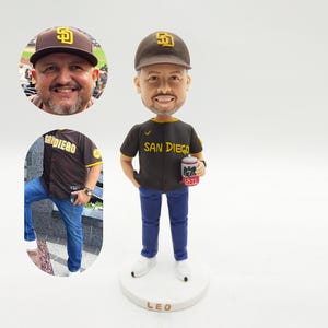 Może przedstawiać: Figurka bobblehead przedstawiająca mężczyznę w brązowej czapce bejsbolowej i czarnej koszulce baseballowej San Diego. Figurka trzyma puszkę i stoi na białej podstawie z napisem "LEO". Dwa zdjęcia wstawione pokazują mężczyznę.