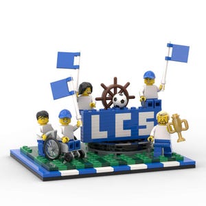 Könnte beinhalten: Ein Lego-Modell einer Fußballmannschaft, die mit blauen Flaggen, einer Trophäe und einem Fußball feiert. Der Name des Teams, "LCS", ist auf einem blau-weißen Schild zu sehen.