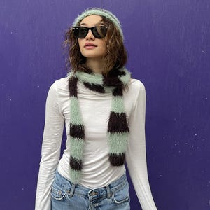 May include: A mint green and brown striped scarf and matching beanie. The scarf is wrapped around the neck, and the beanie sits on the head. The person is wearing black sunglasses, a white long-sleeve shirt, and blue jeans.