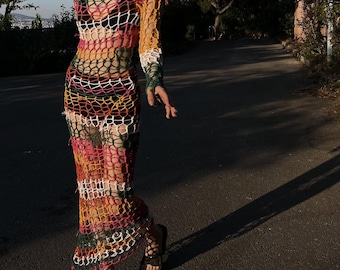 May include: A colorful, open-weave crochet dress with long sleeves. The dress features a mix of yellow, pink, green, and white stripes. The model is wearing black sunglasses and sandals. The dress is a unique, bohemian style.