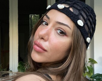 May include: A black crocheted headband with a grid design, adorned with white seashells. The headband is worn on the head. The person has long brown hair and is looking at the camera.