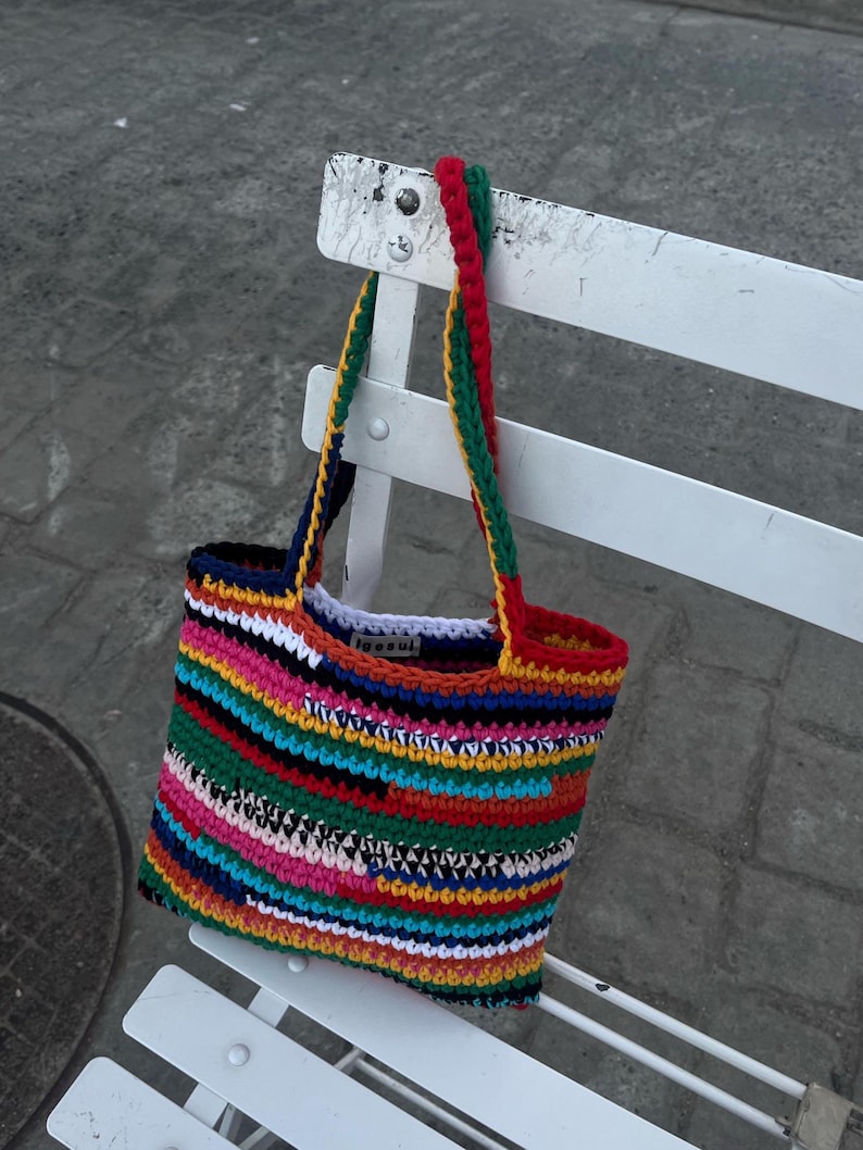 May include: A colourful, handmade crochet tote bag with a rainbow stripe design. The bag has a red, green, and yellow handle. The bag is hanging on a white bench. A unique and colourful accessory.