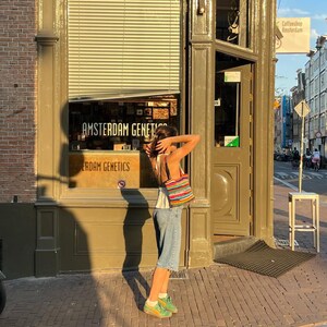 May include: A person stands on a brick pavement outside a shop displaying "Amsterdam Genetics". They wear denim shorts, a white top, and green trainers, carrying a striped bag. The shop is in a building with a brown facade.