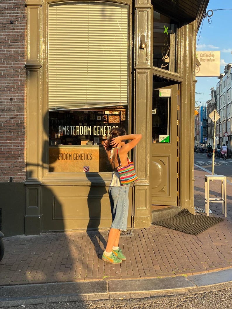 May include: A person stands on a brick pavement outside a shop displaying "Amsterdam Genetics". They wear denim shorts, a white top, and green trainers, carrying a striped bag. The shop is in a building with a brown facade.