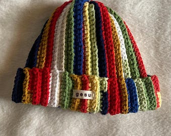May include: A colorful, striped crochet beanie in shades of red, blue, green, yellow, and white. The hat features a folded brim and a small tag with the word "gesu". The beanie is made of a soft, textured yarn.