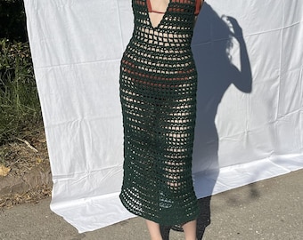 May include: A dark green, open-weave crochet dress with a halter neckline. The dress is knee-length and worn with a rust-colored bikini top. The model is also wearing a wooden bead necklace and dark green sandals.