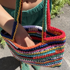 May include: A vibrant, hand-crocheted tote bag featuring a striped design in multiple colours, such as red, orange, yellow, blue, and green. The bag has strong handles and a white interior with the text "igesul".