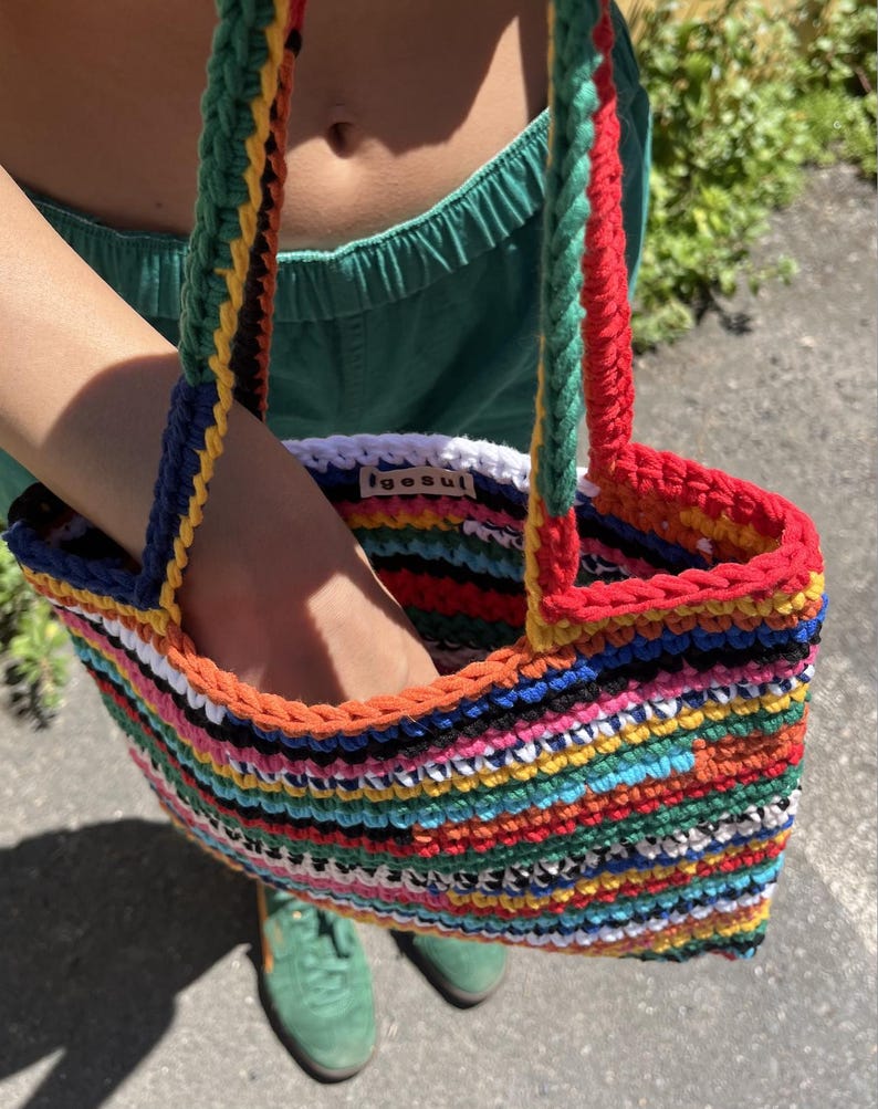 May include: A vibrant, hand-crocheted tote bag featuring a striped design in multiple colours, such as red, orange, yellow, blue, and green. The bag has strong handles and a white interior with the text "igesul".