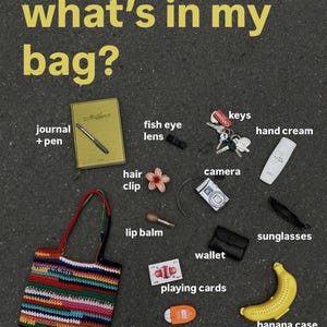 May include: A collection of everyday essentials, including a journal, pen, fish eye lens, keys, hand cream, camera, sunglasses, wallet, playing cards, sunscreen, banana case with banana, and a colourful crocheted bag. The text "what's in my bag?" is at the top.