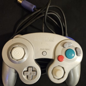 May include: A silver Nintendo GameCube video game controller with a black cord and purple connector. The controller features a central Start/Pause button, a white analog stick, a white C-stick, a green A button, a red B button, and gray X, Y, and D-pad buttons.