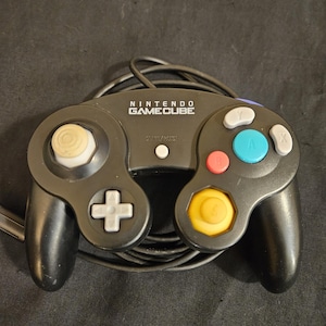 May include: A black Nintendo GameCube video game controller featuring a central Start/Pause button, a white D-pad, a yellow C-stick, a large green A button, a red B button, and grey X and Y buttons, with an attached black wired connection cable.
