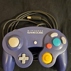 May include: A purple Nintendo GameCube controller with the text "NINTENDO GAMECUBE" at the top. The controller has a white directional pad, a yellow joystick, and colored buttons. A black cable extends from the top.