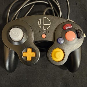 May include: A black wired game controller featuring a white Super Smash Bros. logo, a white analog stick, an orange D-pad, a yellow C-stick, a large orange button, a small red button, a blue button, a black button, and a red Start/Pause button.