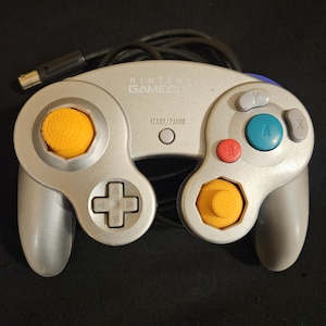 May include: A silver Nintendo GameCube controller featuring a large green A button, a red B button, a gray X button, a gray Y button, two yellow analog sticks, a gray directional pad, and a Start/Pause button with a black wired connector.