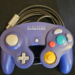 May include: A purple Nintendo GameCube video game controller with a wired connection. The device features a white analog stick, a yellow C-stick, a red B button, a green A button, grey X and Y buttons, a grey D-pad, and a white Start/Pause button.