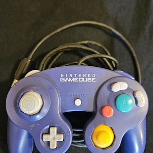 May include: A purple Nintendo GameCube video game controller with a black wired cable. The controller features a central Start/Pause button, a gray directional pad, a large green A button, a red B button, and gray X and Y buttons.