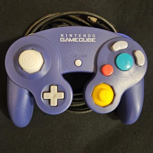 May include: A purple Nintendo GameCube video game controller featuring a white analog stick, a white D-pad, a large green A button, a red B button, gray X and Y buttons, a yellow C-stick, and a Start/Pause button with an attached black cable.