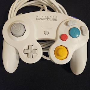 May include: A white Nintendo GameCube controller featuring a gray analog stick, a gray D-pad, a red B button, a teal A button, gray X and Y buttons, a yellow C-stick, and a white Start/Pause button with an attached white cable.