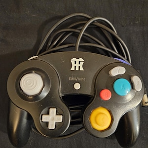 May include: Black video game controller with a white directional pad, gray analog stick, and colored buttons. The controller has a Start/Pause button and a logo. The cord is black and coiled.