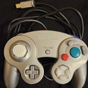 May include: A silver Nintendo GameCube video game controller featuring a central Start/Pause button, a large green A button, a red B button, grey X and Y buttons, a white analog stick, a grey D-pad, and a black wired cable with a connector.