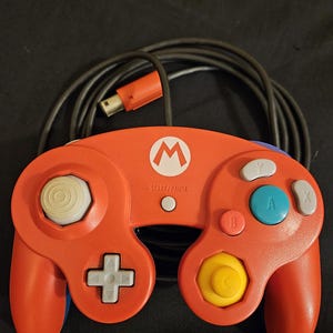 May include: An orange video game controller with a Mario logo. The controller has a white joystick, a yellow directional pad, and blue, green, and gray buttons. A black cord with an orange connector is attached.