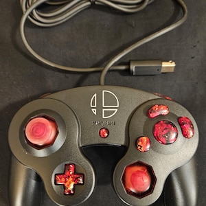 May include: A black wired game controller featuring a white Smash Bros logo and the text START/PAUSE. The controller includes custom red and black marbled resin buttons, a red analog stick, a red C-stick, and a red D-pad with a long black cable.
