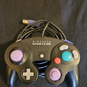 Puede incluir: Un mando de videojuegos Nintendo GameCube negro con un botón central de Start/Pause, un botón A verde, un botón B rojo, botones X e Y grises, una cruceta gris y dos joysticks analógicos morados, con un cable negro y un conector.